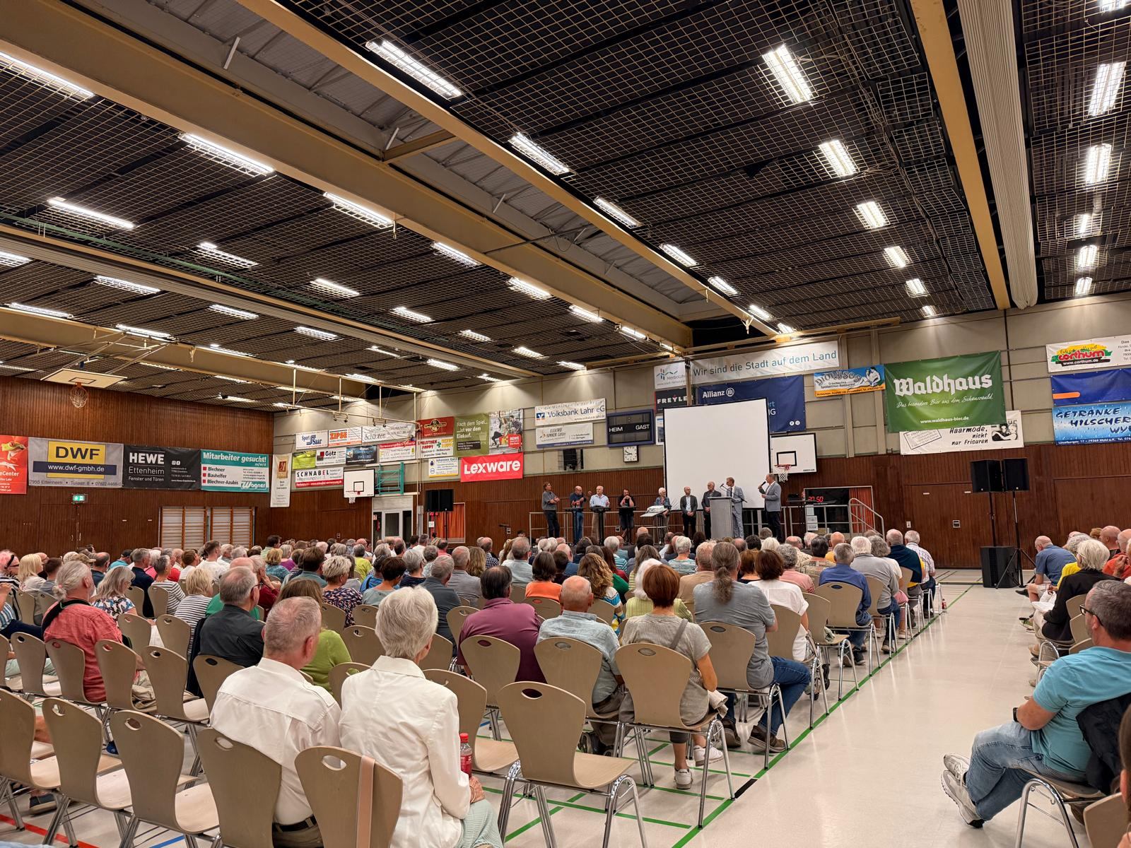 Volle Sporthalle beim Informationsabend, Podium mit Rednerinnen und Rednern vor einer Leinwand
